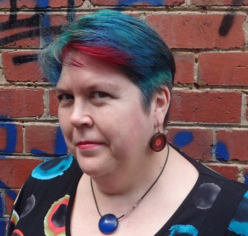 A woman stands in front of a brick wall. She has short red, blue and green hair, and wears a black shirt with red and blue spots. 