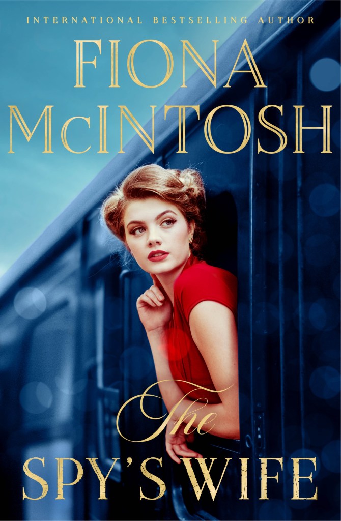 A blonde woman in a red dress on  a train, The Spy's Wife by Fiona McIntosh. The text is in gold. 