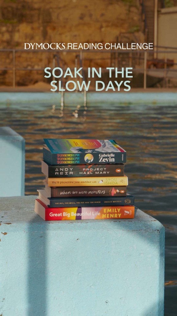 A stack of books on a blue wall next to a pool in sunlight. White text says Dymocks Reading Challenge. Soak in the Slow Days. 